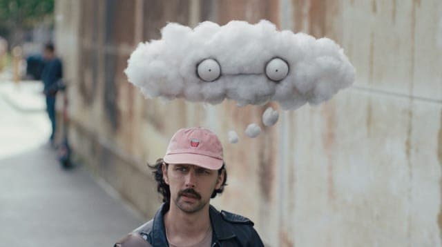 The image shows a man standing on a sidewalk, wearing a pink baseball cap and a dark-colored jacket. He has a mustache and is looking directly at the camera. Above his head, a white cloud with a face drawn on it floats in the air. The cloud has two eyes and a mouth, giving it a whimsical appearance. The man is position