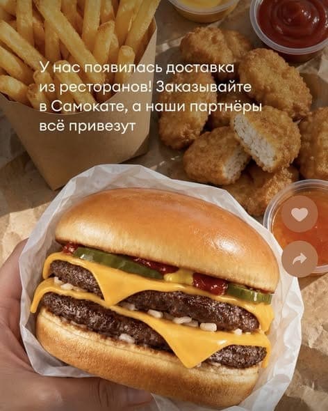 A hand holds a cheeseburger with two beef patties, cheese, pickles and ketchup in paper, with fries, chicken nuggets and dipping sauces on the table behind it. The overlaid Russian text reads "У нас появилась доставка из ресторанов! Заказывайте в Самокате, а наши партнёры всё привезут" ("We now have restaurant delivery
