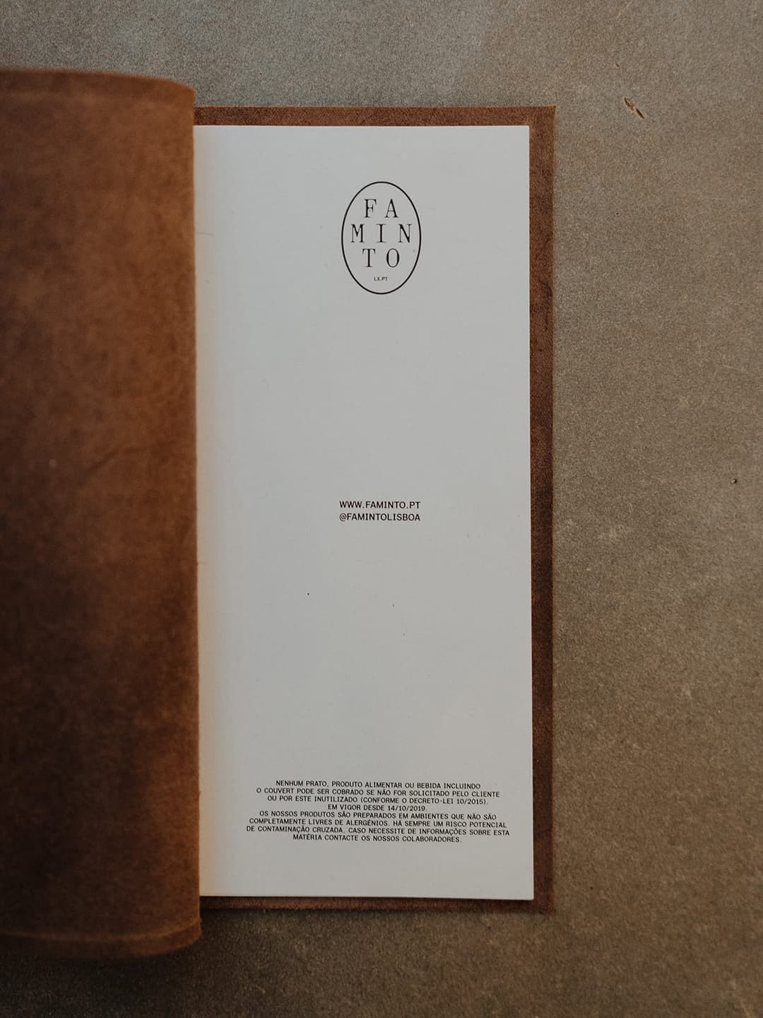 The image shows a book with a brown cover and a white interior page. The book is open, revealing a white interior page with a black logo that reads "FAMINTO" and "LXPT". The page also includes a website address: "www.faminto.pt" and a social media handle: "@famintolisboa". The book is resting on a gray surface.