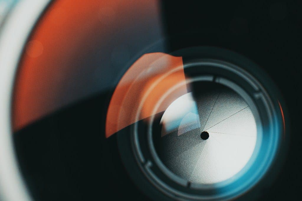 The image shows a close-up view of a camera lens, with a portion of the lens's outer ring visible. The lens is primarily black, with a circular opening in the center. The lens is surrounded by a ring of blue and white, which appears to be a lens flare or light reflection. The background is dark, providing a contrast to