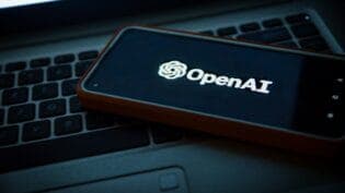 The image shows a black laptop with a reddish-brown or orange case resting on a black keyboard. The laptop screen displays the white text "OpenAI" in a sans-serif font. The laptop and keyboard are positioned on a dark surface, possibly a desk or table.