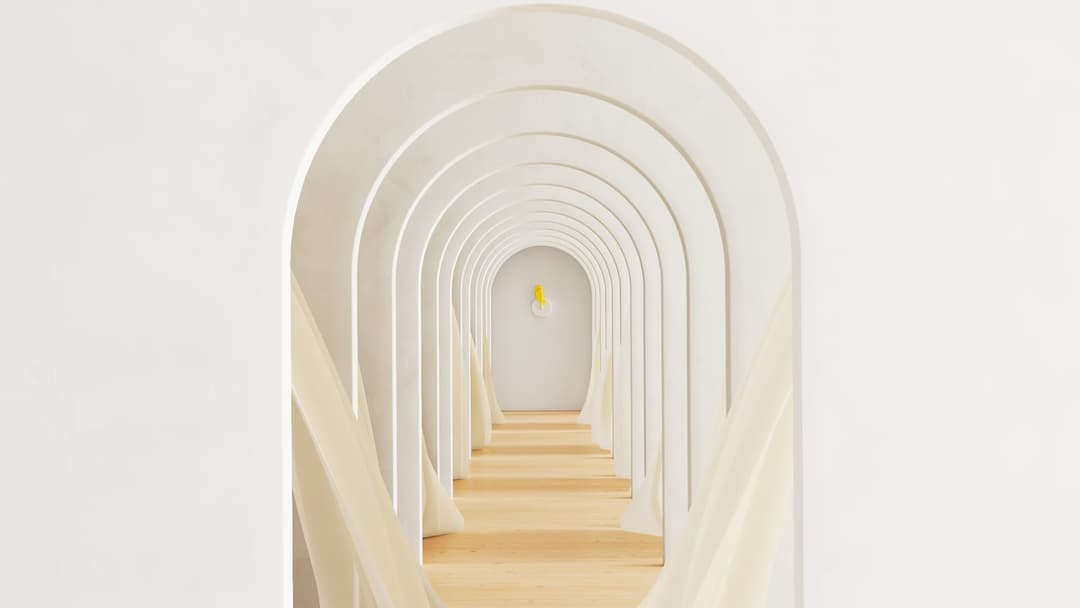 The image presents a serene and minimalist interior scene. Dominating the center of the image is a long, narrow corridor with a series of arches on either side, creating a rhythmic pattern. The arches are painted in a pristine white color, contrasting with the light-colored wooden floor beneath them. The corridor is il