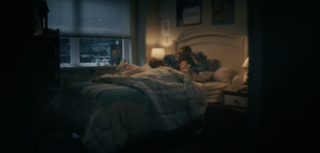 The image shows a dimly lit bedroom with a bed in the center, covered in a blue and white plaid comforter. A person is lying on the bed, possibly reading a book, while another person is sitting up, possibly watching a television. The room is decorated with posters on the wall, and a window with blinds is visible on the