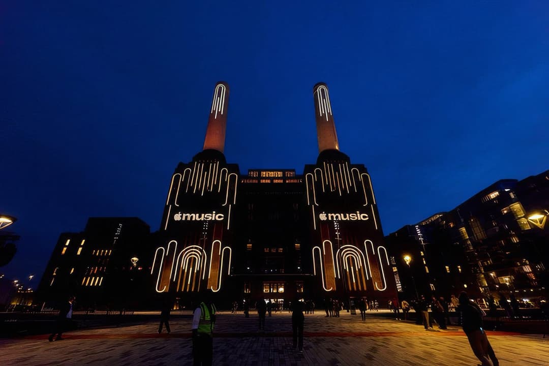 The image captures the iconic Battersea Power Station in London, England, at night. The power station is illuminated by a warm, orange glow, contrasting with the dark blue sky. The two towering smokestacks, each adorned with a white, stylized "Apple Music" logo, are the central focus of the image. The power station's f