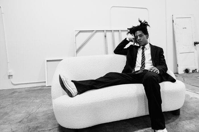 In this black and white photograph, a young man is seated on a white couch in a room with a concrete floor. He is dressed in a suit and tie, with his hair styled in dreadlocks. His left foot is resting on the armrest of the couch, while his right hand is resting on his head. The room features a white door on the right