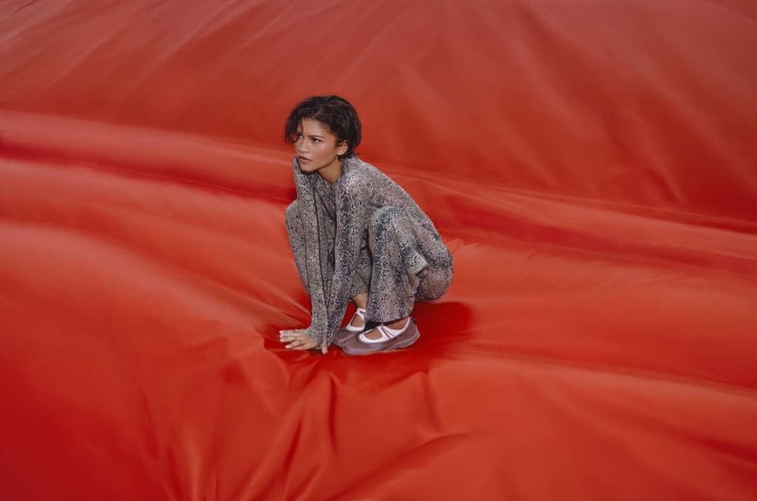 The image shows a young woman crouching on a large, red, fabric-like surface. She is wearing a gray, patterned, long-sleeved top and matching pants, along with white sneakers. Her gaze is directed off to the side, and her expression is neutral. The red surface appears to be a large, inflated object, possibly a balloon