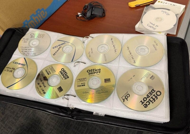 The image shows a black binder with a white insert, neatly arranged with eight CD-ROMs. The binder is open, revealing the CDs within. The CDs are organized in two rows of four, with the top row slightly offset from the bottom row. The CD-ROMs are predominantly gold or beige in color, with some featuring blue and white